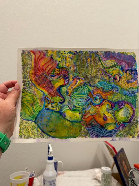 A colorful abstract mixed media painting with swirling watercolor, ink, and pencil marks, held up by the artist against a neutral wall. The paper edges curl naturally.
