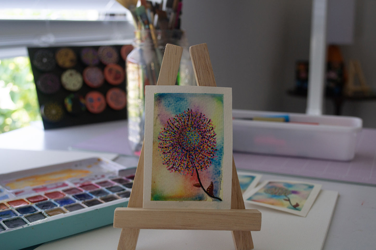 Small watercolor painting of a colorful flower on a wooden easel with art supplies in the background.