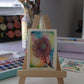 Small watercolor painting of a colorful flower on a wooden easel with art supplies in the background.