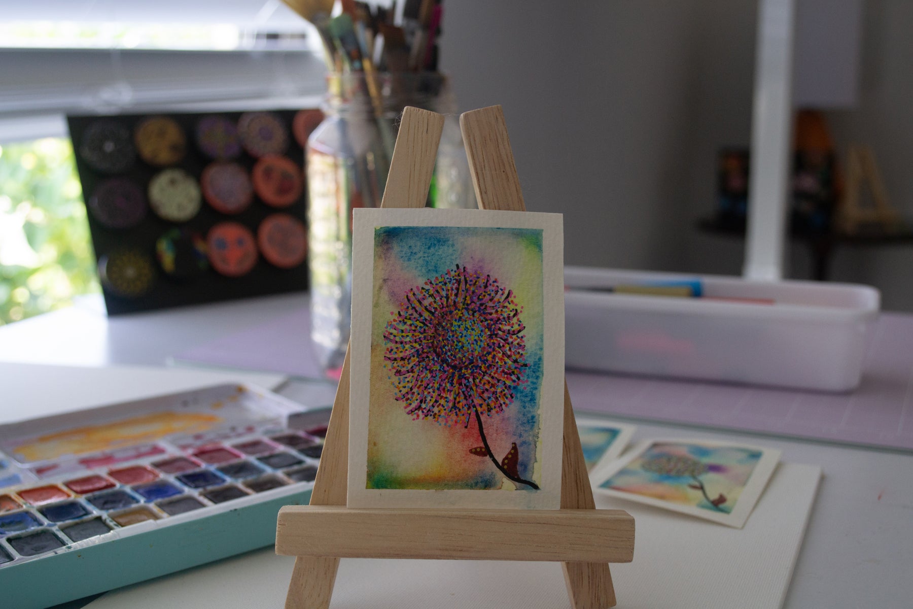 Small watercolor painting of a colorful flower on a wooden easel with art supplies in the background.